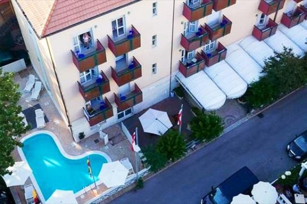 Pohľad zhora na budovu s bazénom a balkónmi vedľa úzkej cesty so zaparkovanými autami.