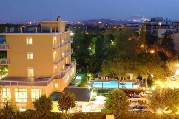 Večerný pohľad na osvetlený hotel s bazénom obklopený stromami a mestskou krajinou v pozadí.  