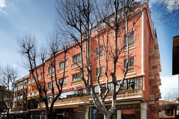 Štvorposchodová budova s ružovou fasádou a balkónmi, pred ktorou parkujú autá a rastie niekoľko holých stromov.