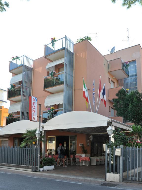 Trojposchodová budova s balkónmi a strešnou záhradou, s vlajkami na fasáde nad vstupom.