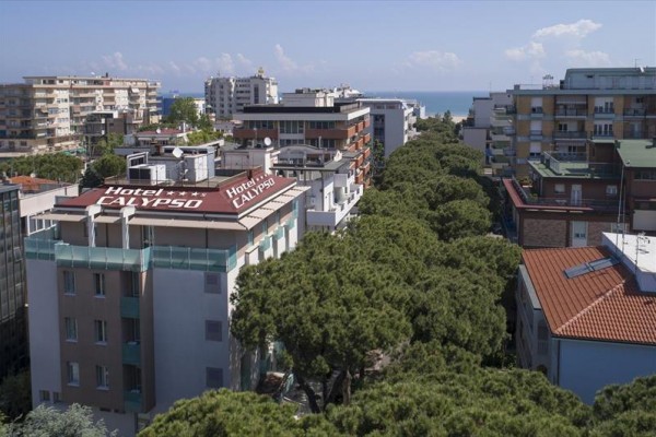 Pohľad na mestskú štvrť s hotelom Calypso a zelenými stromami, v pozadí je vidieť more.  