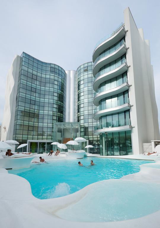 Moderný hotel s bazénom, okolo ktorého sú rozmiestnené lehátka a slnečníky.