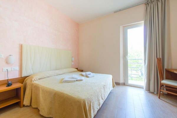 Hotelová izba s manželskou posteľou a otvorenými dverami na balkón.