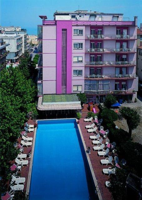 Ružový hotel so štyrmi poschodiami a vonkajším bazénom obklopeným lehátkami.