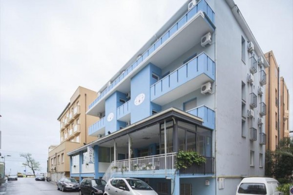Moderná modro-biela trojposchodová budova s balkónmi a zaparkovanými autami pred vchodom.