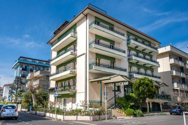 Viacposchodová budova s balkónmi a zeleňou pred vchodom na mestskej ulici.
