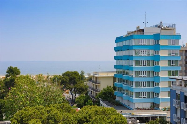 Modro-biely viacposchodový hotel pri mori, obklopený zeleňou.