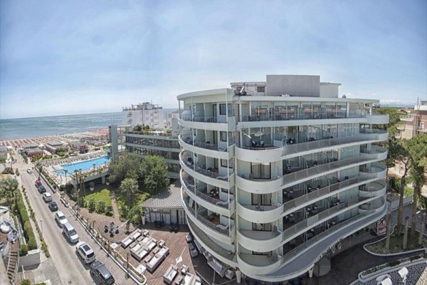 Moderný hotel pri mori s balkónmi a veľkým bazénom vedľa pláže.