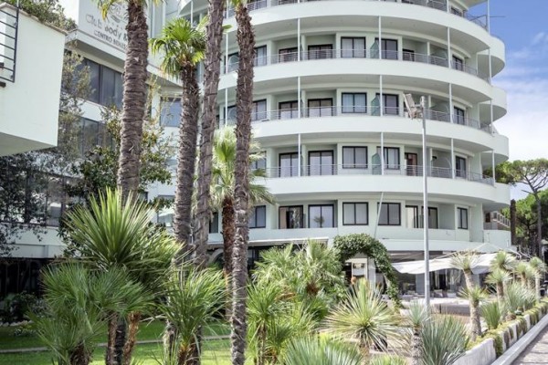 Moderná zaoblená budova s balkónmi obklopená palmami a zeleňou.