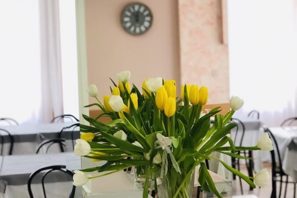 Kytica žltých a bielych tulipánov na stole v miestnosti s okrúhlymi stolmi a hodinami na stene.