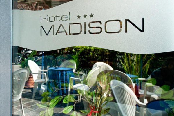 Logo hotela Madison na sklených dverách s odrazom stoličiek a rastlín.
