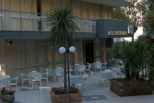 Vonkajšia terasa s bielymi kovovými stolmi a stoličkami obklopená kvetináčmi a palmami pred hotelom.