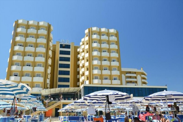 Veľký hotel s balkónmi a modrobielymi plážovými slnečníkmi na piesočnej pláži.