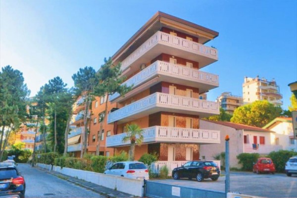 Štvorposchodová obytná budova s balkónmi obklopená stromami a autami na ulici.