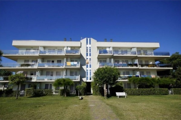 Štvorposchodová obytná budova s balkónmi a záhradou pred ňou na pozadí modrej oblohy.