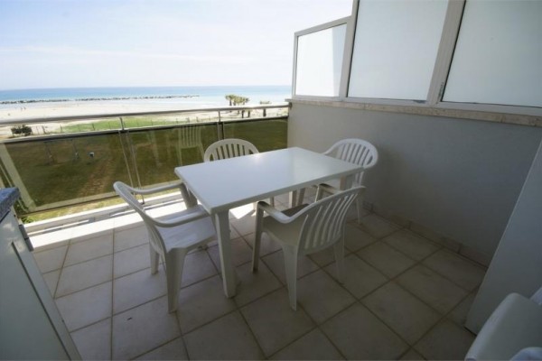 Balkón s výhľadom na pláž a more, vybavený štyrmi bielymi plastovými stoličkami a stolom.