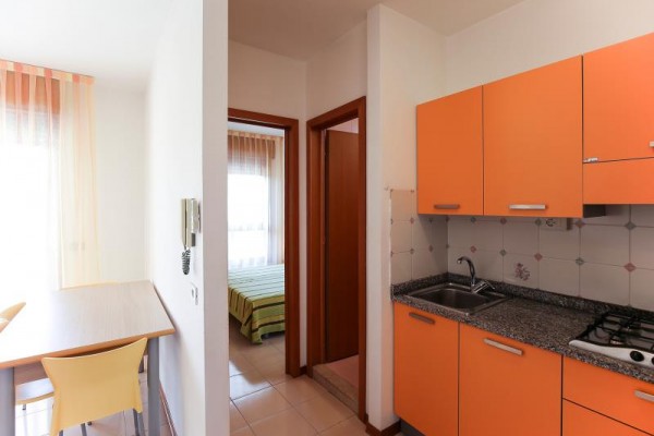 Moderná kuchyňa s oranžovými skrinkami vedľa jedálenského kúta a dverí do spálne.