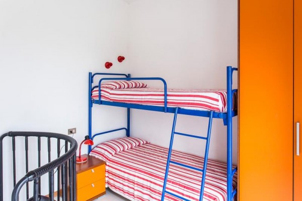 Poschodová posteľ s červeno-bielymi pruhovanými obliečkami, vedľa je oranžová skriňa a nočný stolík s lampou.