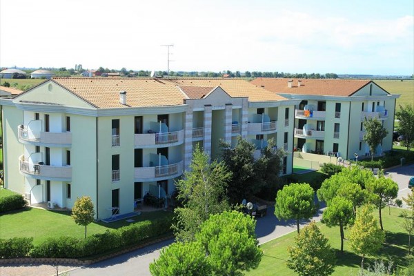 Trojposchodová bytová budova s balkónmi obklopená zeleňou a stromami.