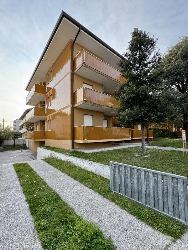 Moderný bytový dom s tromi podlažiami a balkónmi, obklopený zeleňou a stromom.