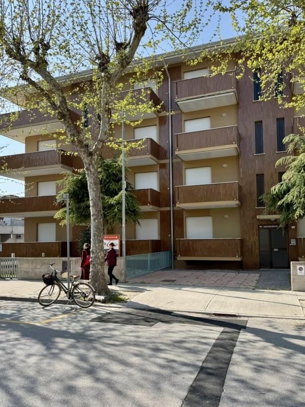 Viacposchodová budova s balkónmi a stromom v popredí.