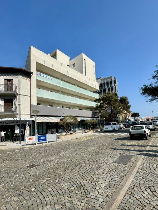 Moderná budova s balkónmi na rohu dláždenej ulice počas slnečného dňa.