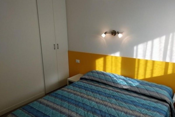 Spálňa s manželskou posteľou, modrošedou prikrývkou, žltým panelom na stene a bielou skriňou.