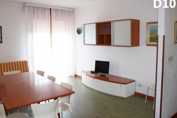 Jednoducho zariadená obývačka s televíziou, stolom a stoličkami pri veľkom okne s bielymi závesmi.