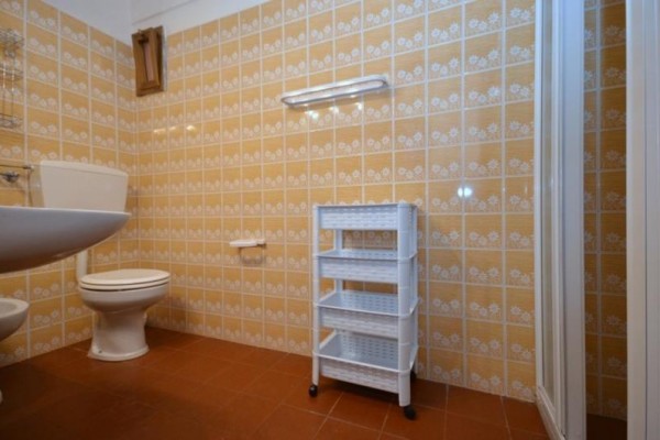 Kúpeľňa s oranžovými obkladmi, toaletou, umývadlom a bielym plastovým regálom.