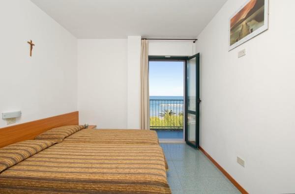 Spálňa s manželskou posteľou a otvorenými dverami vedúcimi na balkón s výhľadom na more.